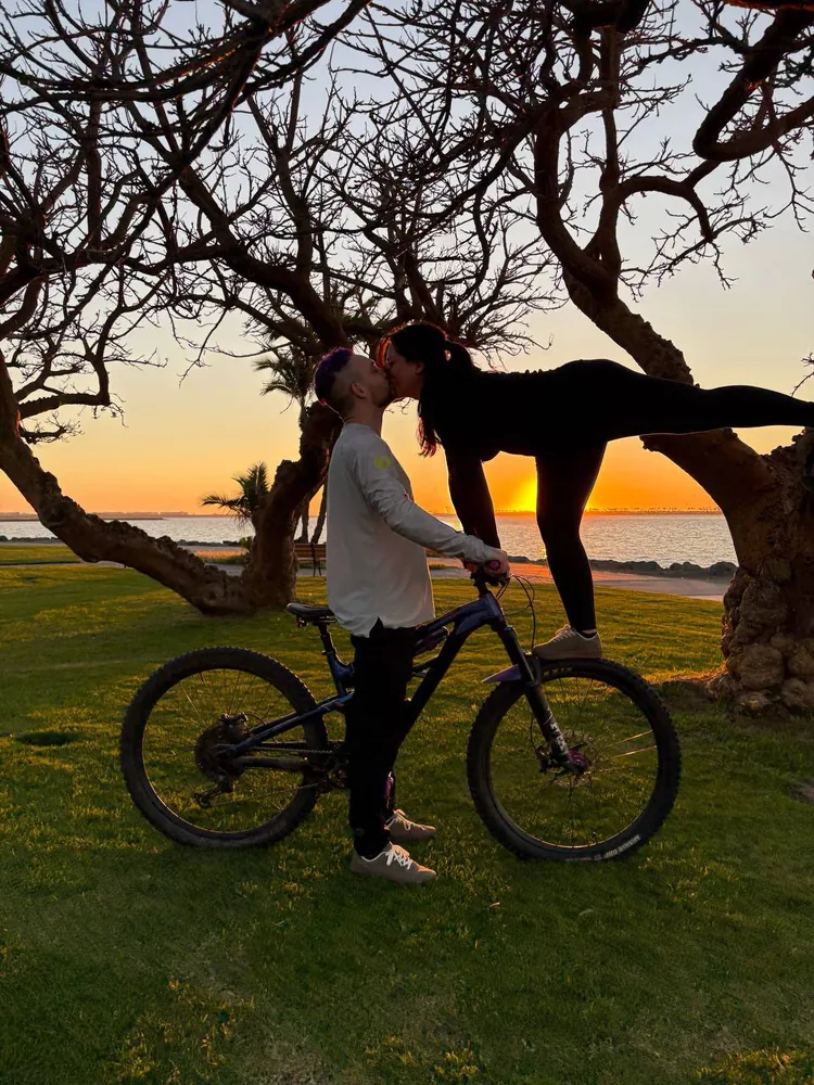 Saray standing on a tree branch to kiss Jeremy, who is straddling his bike, as the sun sets over the bay.