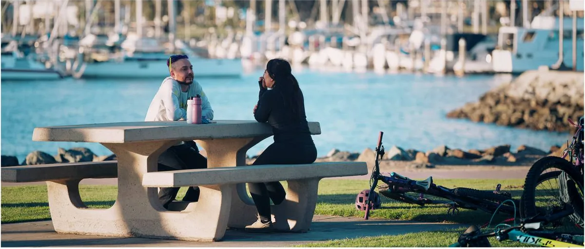 Saray and Jeremy talking across a picnic table at the marina, their bikes lying on the grass beside them.